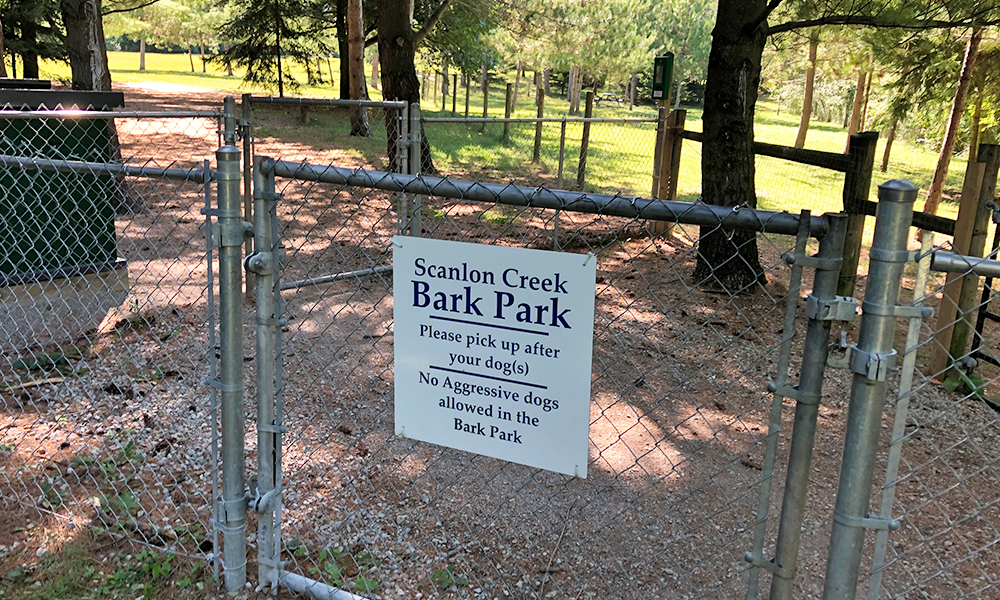 The entrance to the Scanlon Creek Bark Park is marked by a fenced gate, offering a glimpse of trees in the background and the natural setting for dogs to play and explore.