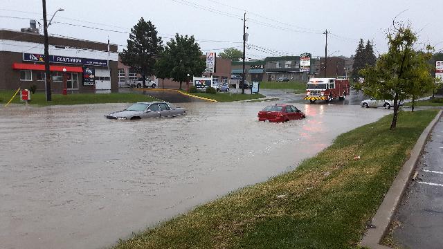 Two cars are partially submerged on a significantly flooded street in Aurora, Ontario after a heavy rain event. A firetruck is on scene providing emergency services.