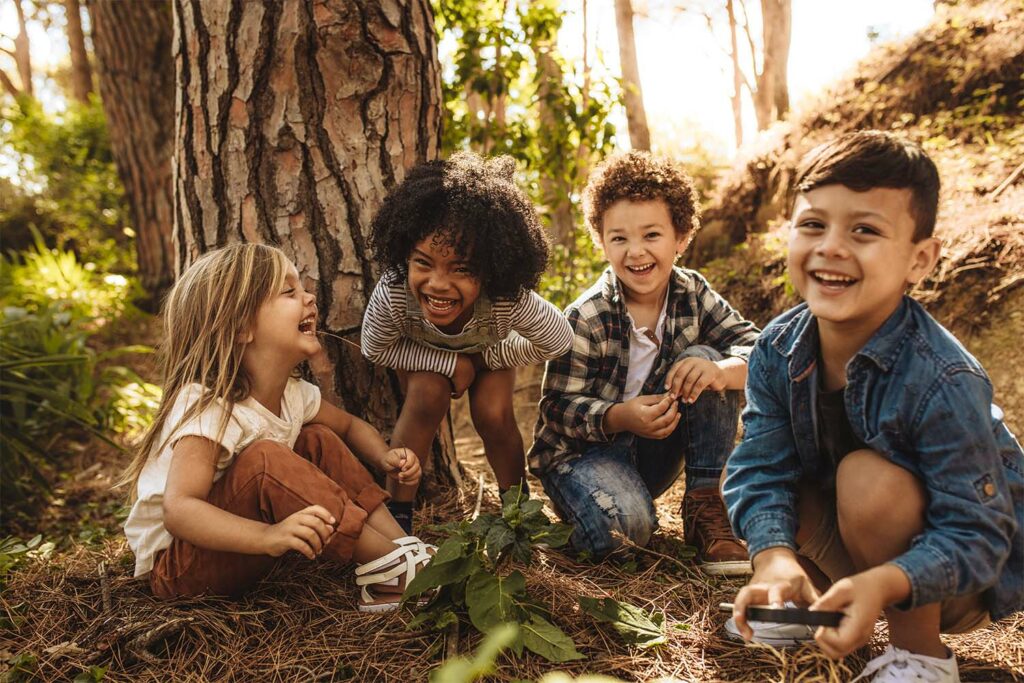 On a bright summer day, four children gather huddled together beside a large tree in the forest. One child holds a magnifier in his hand, while all four children laugh and enjoy themselves in the natural surroundings.