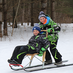 During March Break camp, two adolescents enjoy a winter adventure as one comfortably sits on the kick sled while the other stands just behind him after energetically propelling the sled forward.