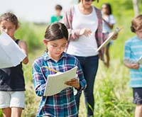 A multi-ethnic group of elementary school children are outdoors exploring nature. The children are busy writing on their notepads while walking with a teacher.