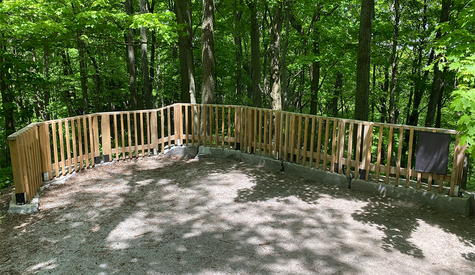 A waist-high wooden railing is securely mounted atop of a row of sturdy armor stones, encircling one side of a viewing platform at the Thornton Bales Conservation Area. Lush green forest is visible in the background.