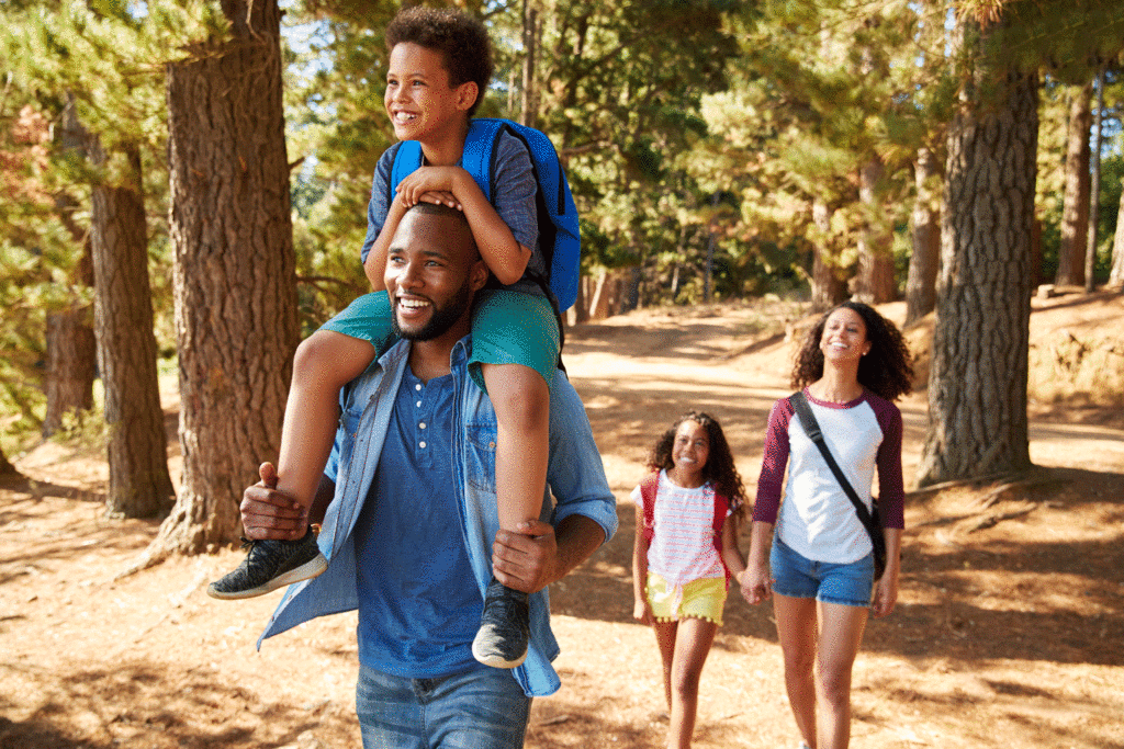 A family leisurely strolls through the forest, creating a heartwarming scene. A young boy sits on his dad's shoulders, his face beaming with joy, while a young girl holds her mom's hand, captivated by the enchanted surroundings.