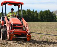 Conservation Authority staff operating a red tractor, used to aid in the planting of several thousand tree seedlings. Several neatly arranged rows of small trees are visible in the expansive background, with a large stand of dark green trees seen at the back of the property.