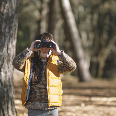 On an autumn day, a child donned in a yellow vest, stands beside a tree, peering through binoculars with curiosity.