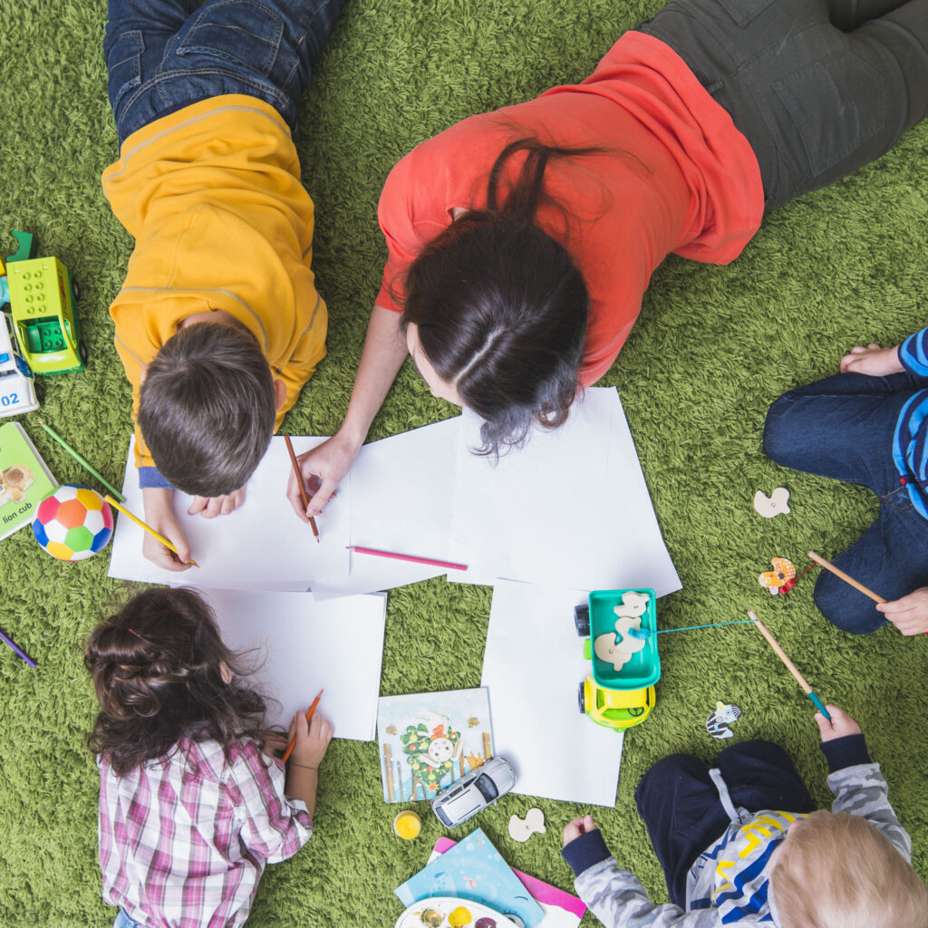 Young children lay on their bellies and immerse themselves in a colouring activity on a vibrant green carpet. Books, toy cars, puzzle pieces and a ball are scattered across the play area.