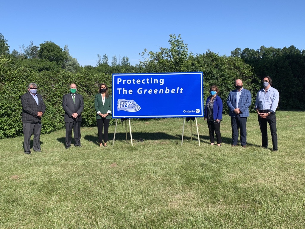 ^ people standing on either side of a sign that says 'Protecting the Greenbelt' for the Maple Lake Estate - Greenbelt Announcement. It's a sunny summers day in a grassy field surrounded by trees.