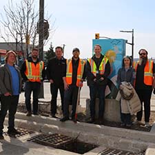 Jacob Reid, and members of the Low Impact Development Municipal Inspection and Maintenance Working Group, wear safety vests and gather on a corner boulevard. In the foreground, a stormwater road drain is open.