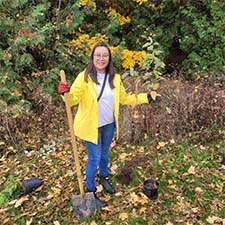 In a grassy area near towering trees, a volunteer clad in a yellow jacket, blue jeans and red gloves, stands with a shovel in hand, poised to plant a tree.